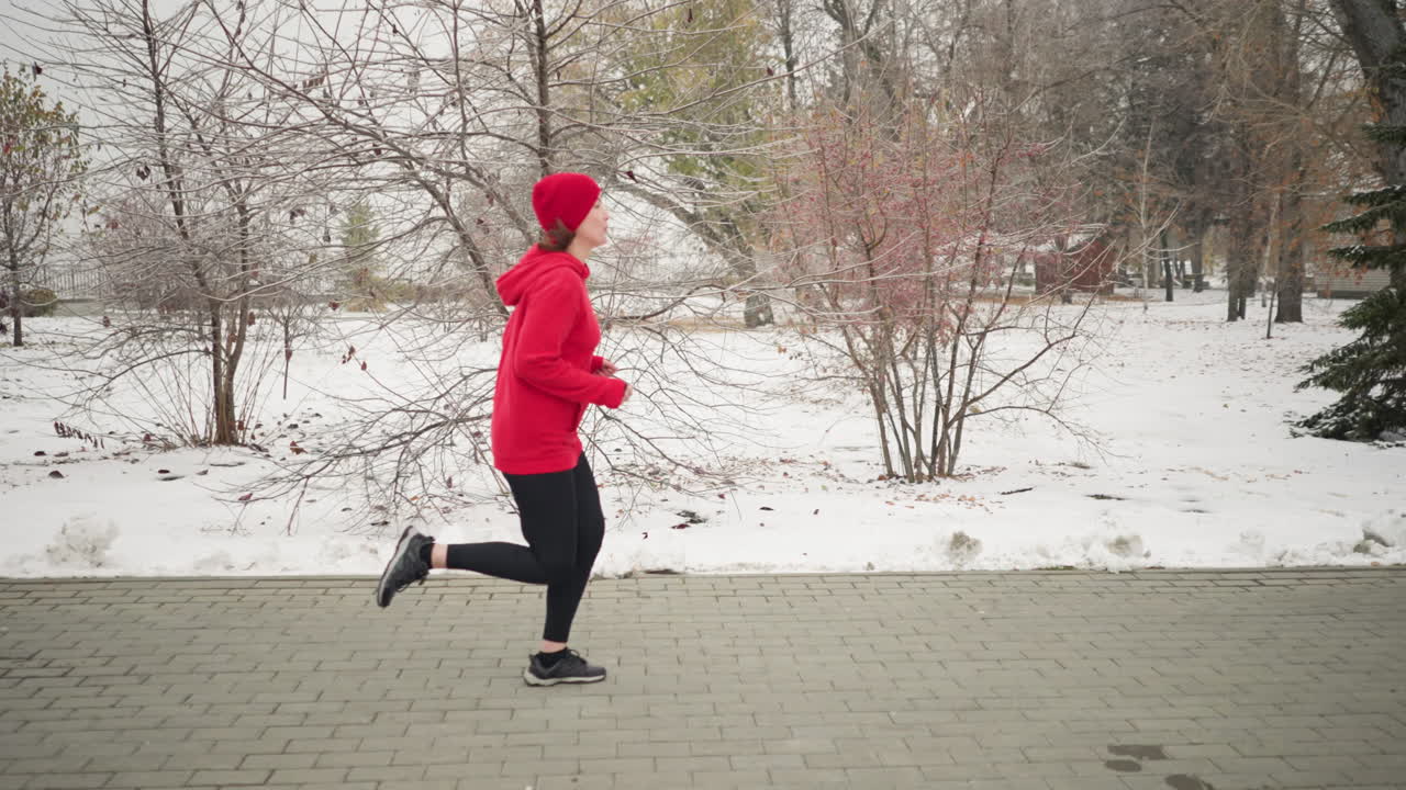 Side view of a woman wearing red beanie and hoodie jogging outdoors during winter surrounded by snow-covered trees, serene park pathway, evergreen pines, and foggy atmosphere