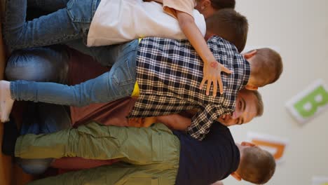 Vertical-video-of-children-running-up-to-their-teacher,-a-man-with-gray-hair-in-a-purple-T-shirt,-and-hugging-him-in-a-club-room-to-prepare-children-for-school