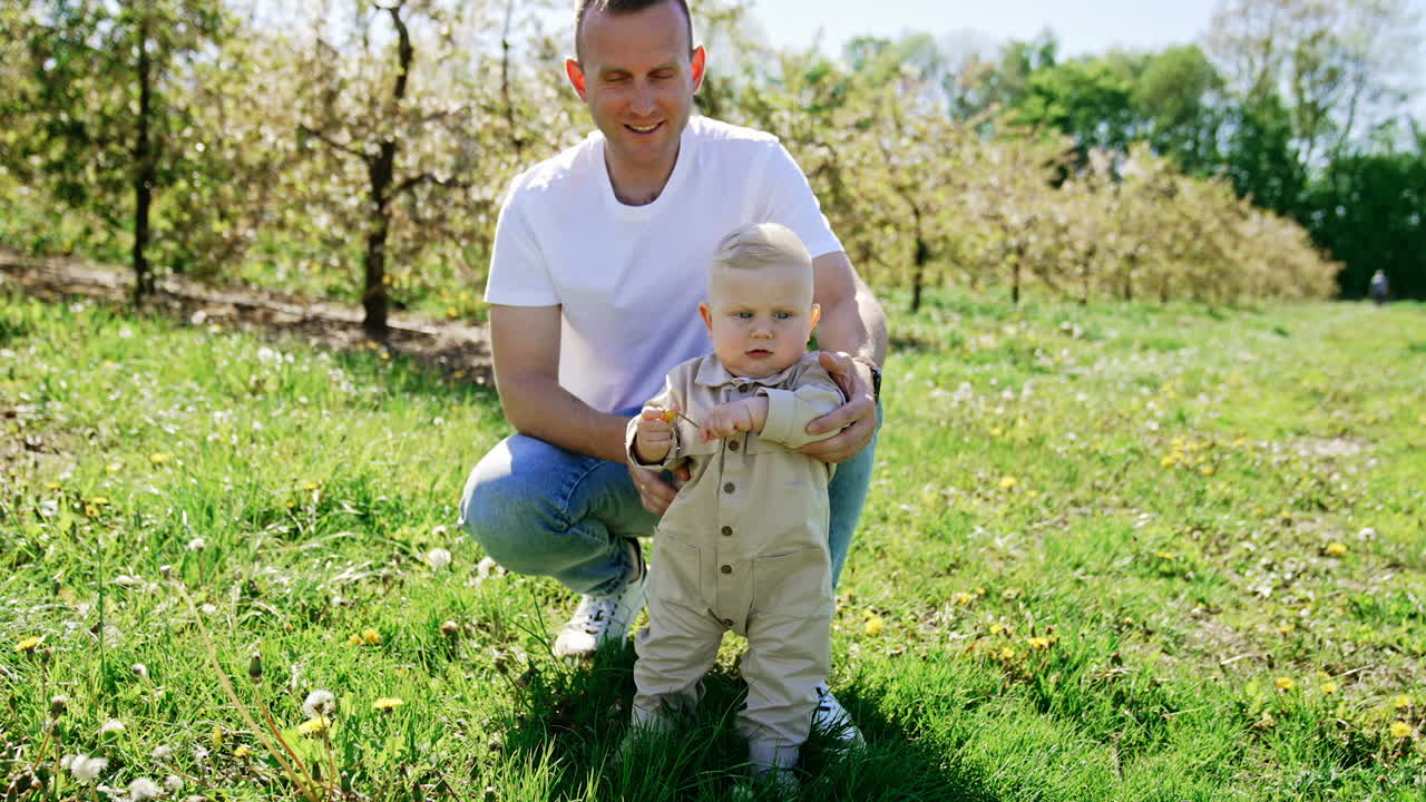 Caucasian man wearing white shirt and jeans sits squatted by his little son standing beside. Lovely baby in beige robe holding a flower. Beautiful sunny spring day in nature.