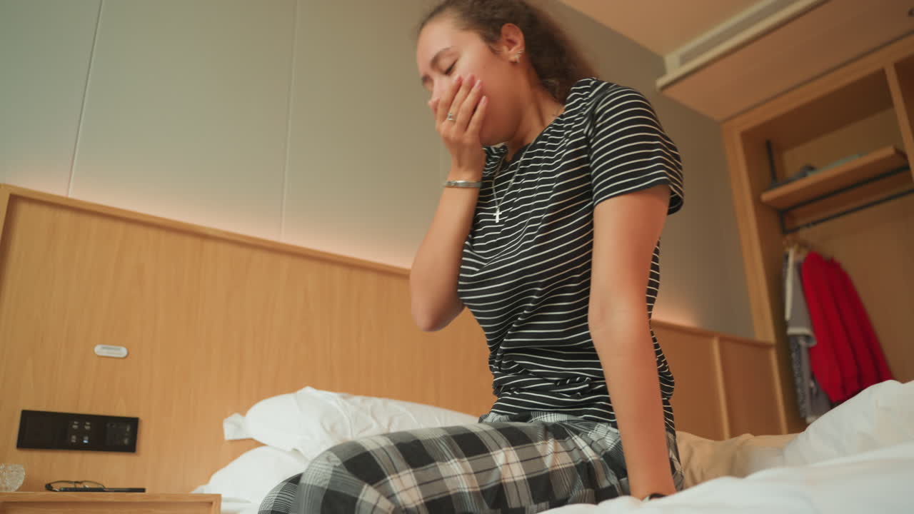 Donna caucasica assonnata sul letto d'albergo che si stira, lenta routine mattutina in una stanza moderna e compatta, vestiti a quadri, lenzuola bianche, testiera in legno, illuminazione soffusa, atmosfera tranquilla da viaggiatrice solitaria