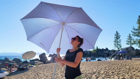Frau-Dreht-Regenschirm-Mit-Sonnenbrille,-Die-An-Einem-Sonnigen-Tag-Am-Strand-In-Lake-Tahoe,-USA,-Steht