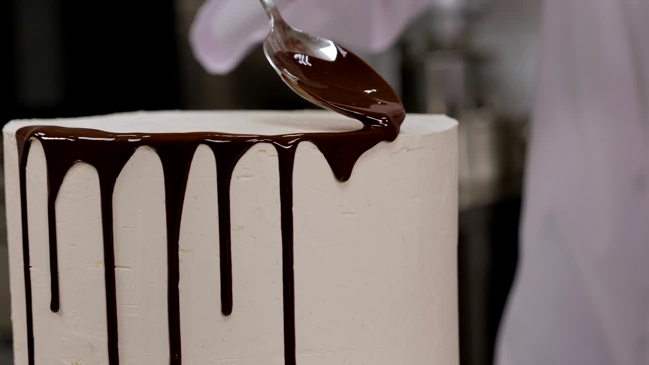 un chef de pastelería vertiendo chocolate líquido de una cuchara en un pastel de esponja de crema blanca