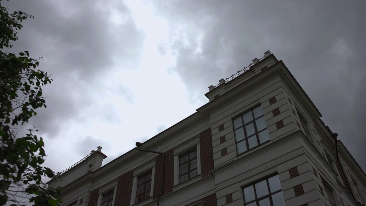 Un moderno edificio urbano con architettura in cemento e vetro si erge sotto un cielo nuvoloso, ripreso da una prospettiva fissa che guarda verso l'alto la struttura. La scena ha un'atmosfera neutra.