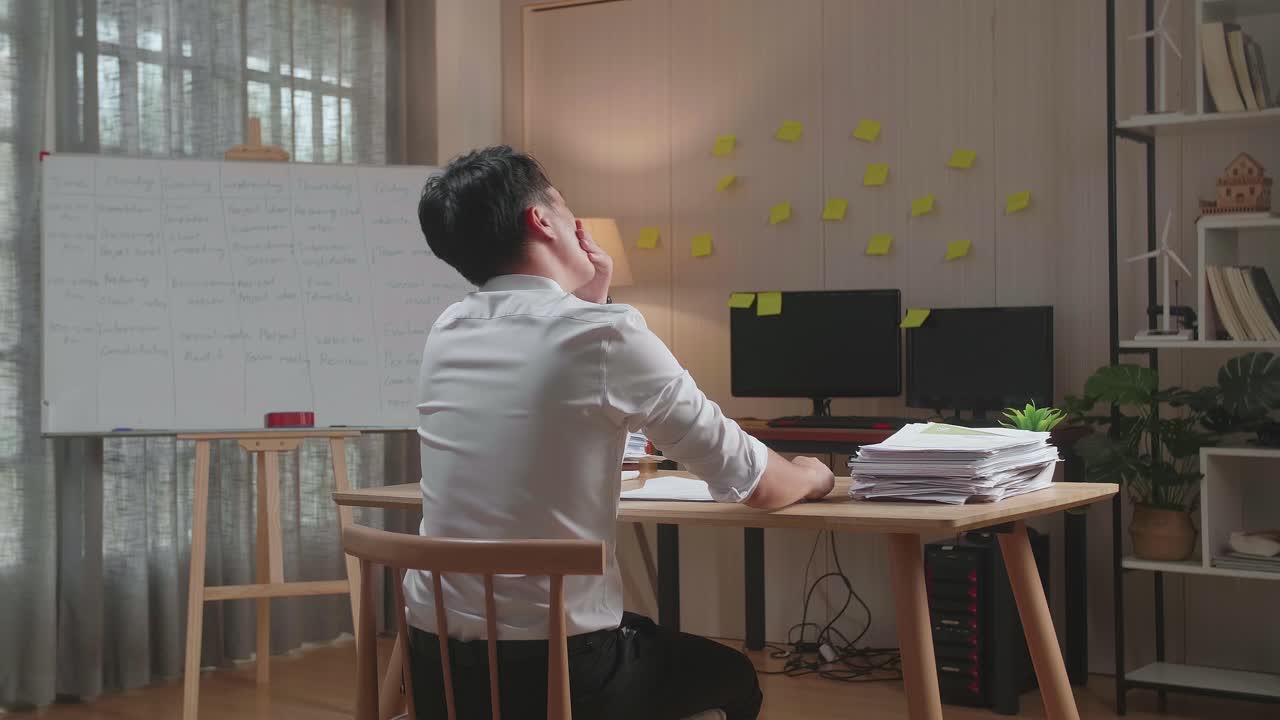 vista trasera de un hombre asiático bostezando antes de dormir mientras trabaja duro con documentos en la oficina