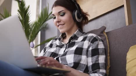 shooting from below of a charming brunette sitting on a sofa. girl in headphones typing on laptop, smiling. communication