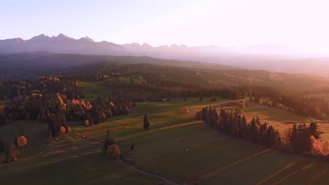 Aerial-shot-of-mountains,-colorful-autumn-trees,-huge-green-fields-and-sunset