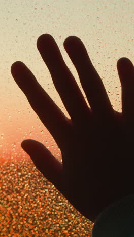 hand silhouette on window covered with raindrops closeup. palm touches wet glass against bright red sunset sun on rainy day. melancholic mood