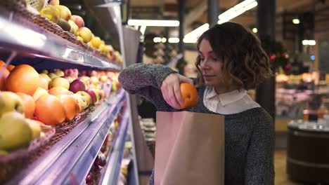 cute girl buys fresh oranges in the market. beautiful young woman stands in front the shelf and puts the oranges to a brown paper bag, she is pleased with the choice