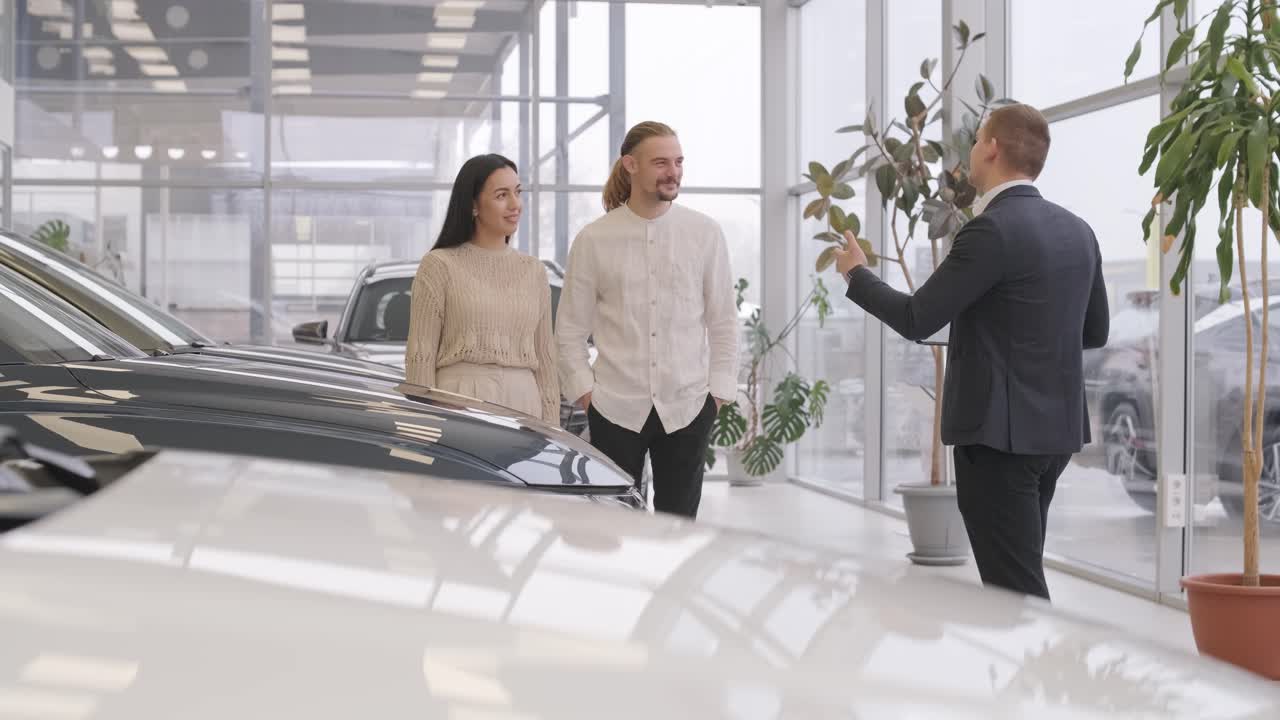 Young car salesman showing to young couple new automobile at dealership salon.