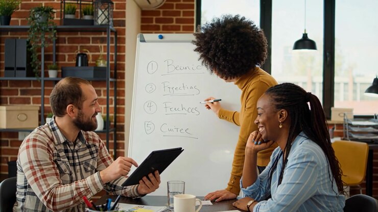 Pointing man in plaid shirt showing tablet, team writing numbered list on flip chart in office