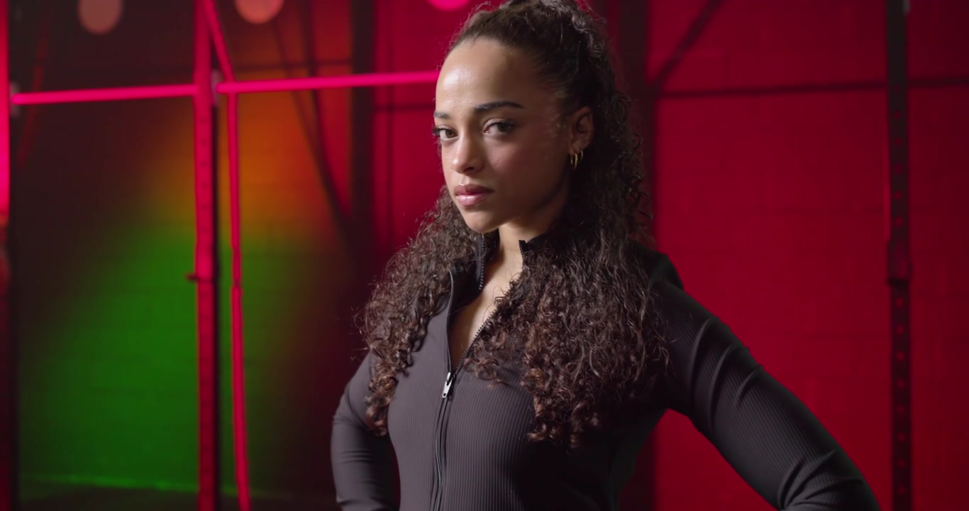 Donna afroamericana in piedi con le mani sui fianchi, con una giacca da palestra, che respira mentre la telecamera si alza, riscaldandosi.