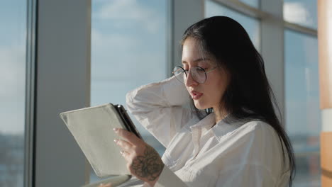 beautiful young woman in white shirt and glasses gently brushes hair back while holding tablet, standing near window with soft sunlight and urban view reflecting on glass surface