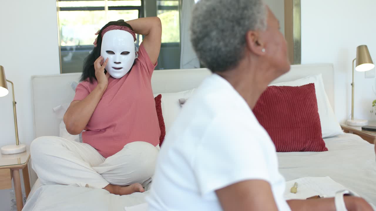 Ældre afroamerikansk kvinde i lyserød top ligger i seng, påfører en hvid ansigtsmaske til hudpleje og holder en telefon.