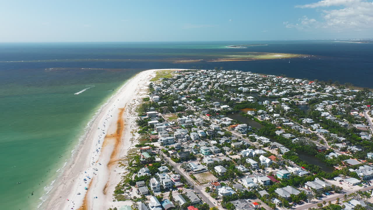 Turquoise waters meet the sandy shoreline of Anna Maria Island, where a coastal town spreads along the beach under a bright summer sky
