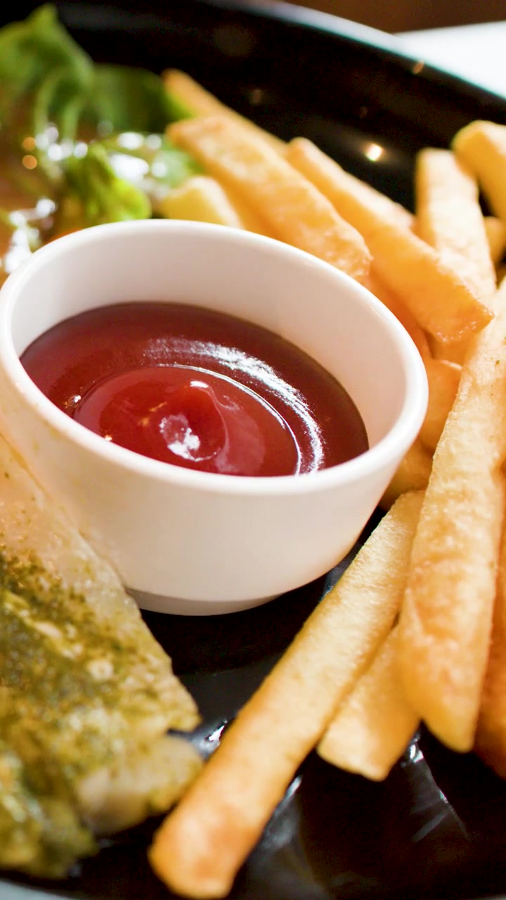 Primo piano di una persona che taglia pesce alla griglia con salsa al pesto, servito con patatine fritte e ketchup