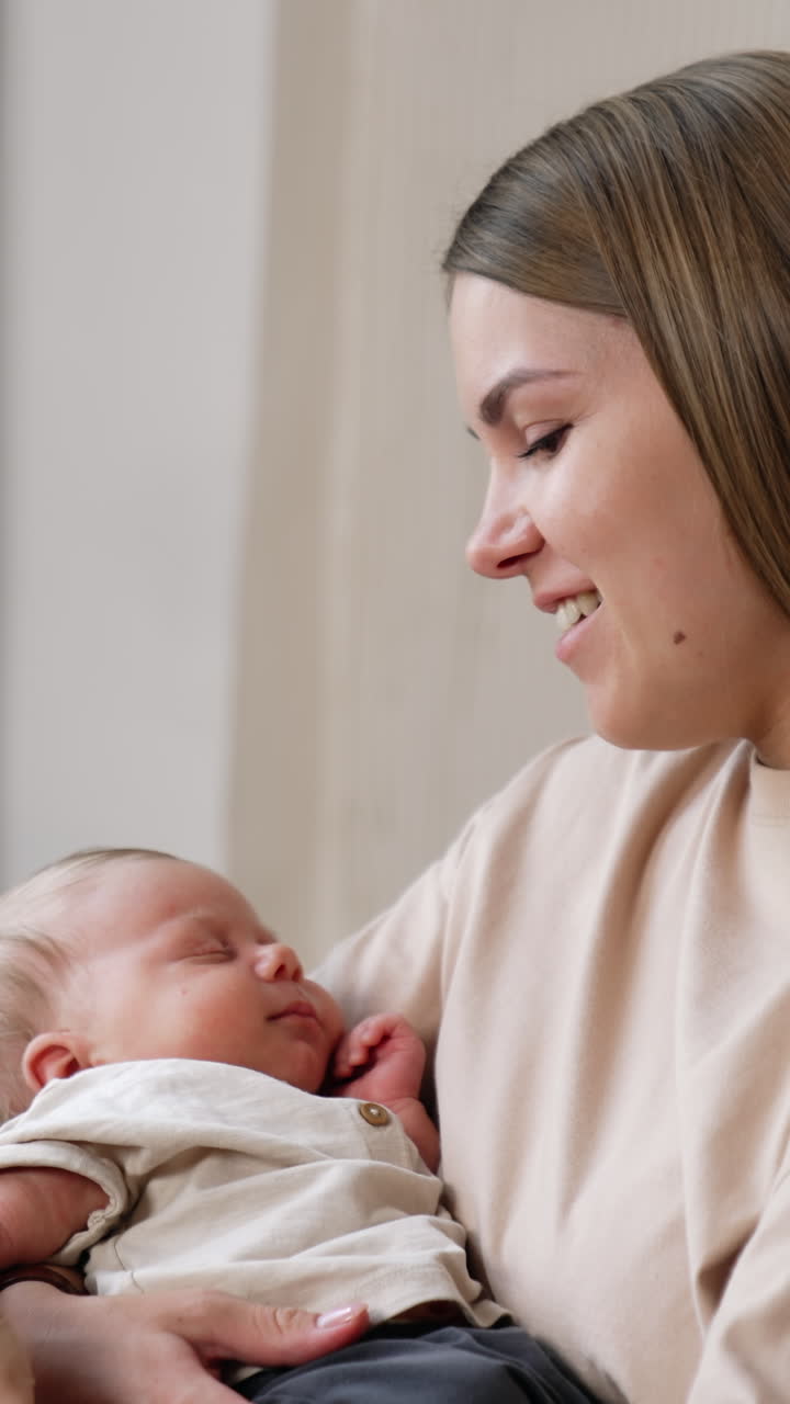 Happy smiling long-haired Caucasian woman waving her infant in arms sitting on a sofa. Mother strokes her child's arms with love. Vertical video