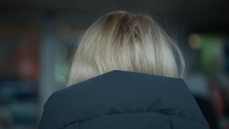 황금색 머리카락을 가진 여자가 후디를 입고, 희미하게 조명된 공간에서 고, 흐릿한 인물이 배경에서 움직여, 부드러운 조명으로 기분 좋은 분위기를 만니다.