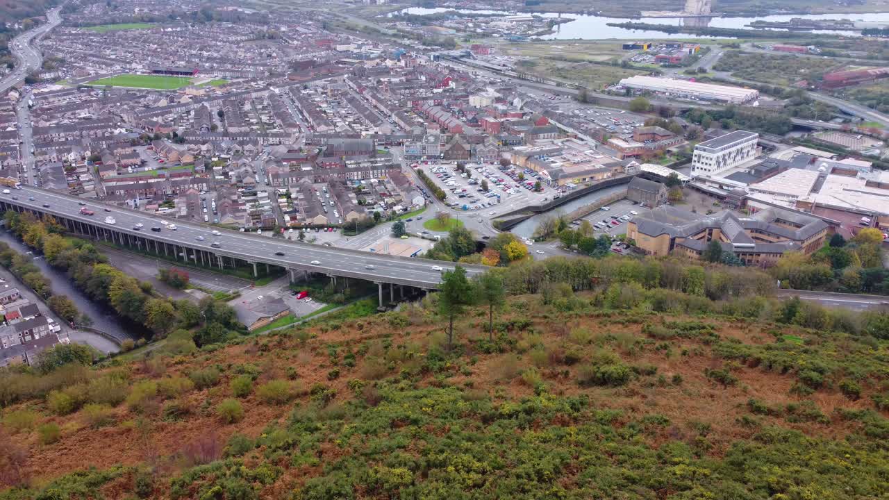 Zijwaarts luchtbeeld van de stad Port Talbot met de M4 snelweg met gestaag verkeer van auto's, busjes en vrachtwagens. Rijen huizen en een lokaal theatergebouw. 4K.