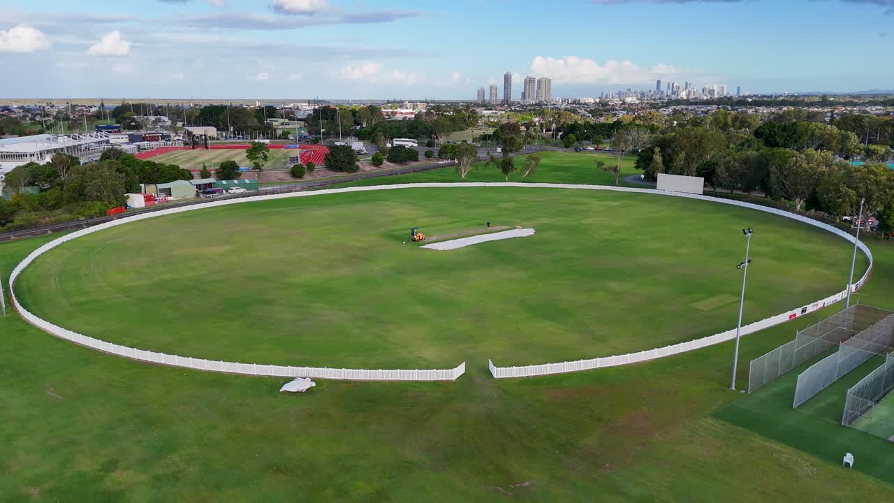Drone footage captures a cricket field in Gold Coast, Australia, under bright daylight, showcasing the surrounding urban landscape