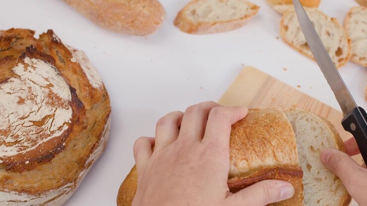 Overhead shot of hands using a knife to slice crusty bread on a wooden board.