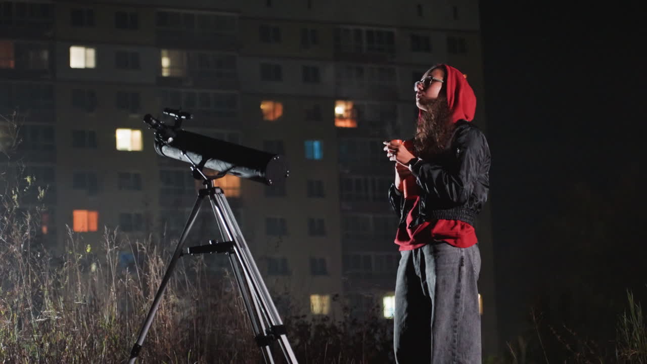 notte di osservazione delle stelle con telescopio sul tetto, un astronomo amatoriale regola l'oculare mentre le finestre della città si illuminano, sciarpa rossa e cappotto pesante, treppiede appoggiato al condominio, smartphone in mano, sguardo rivolto verso l'alto