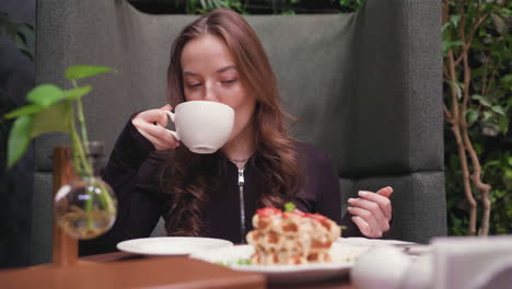 lady enjoys peaceful moment sipping latte with closed eyes, seated at cozy booth beside decorative glass vase with plant and plate of waffles, surrounded by lush greenery and warm tones