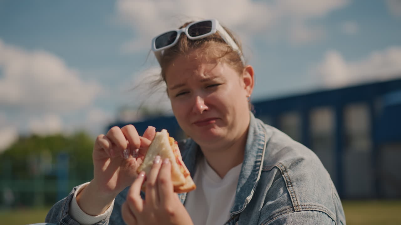 Moment der Essensbewertung. Sandwich-Verkostung im Freien. Ausdrucksstarke Frau probiert Sandwich an einem hellen Sonnentag. Weibliche Food-Enthusiastin genießt Schicht-Sandwich im Park mit Sonnenbrille und Jeans.