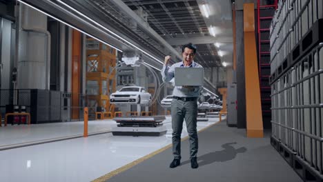 full body of an asian male professional worker standing with his laptop in ev car factory, high-tech industrial facility, he raises his fist up with screaming goal