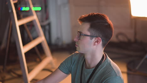 person focused on adjusting video equipment in tv studio, seated and working with control panel. studio lighting visible in background while adjusting settings on monitor and controls