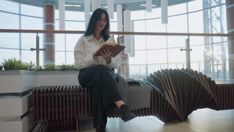 novelist seated on uniquely designed wooden bench in modern mall flips page while reading book, surrounded by glass railing, stylish architecture, and soft daylight highlighting calm, focused