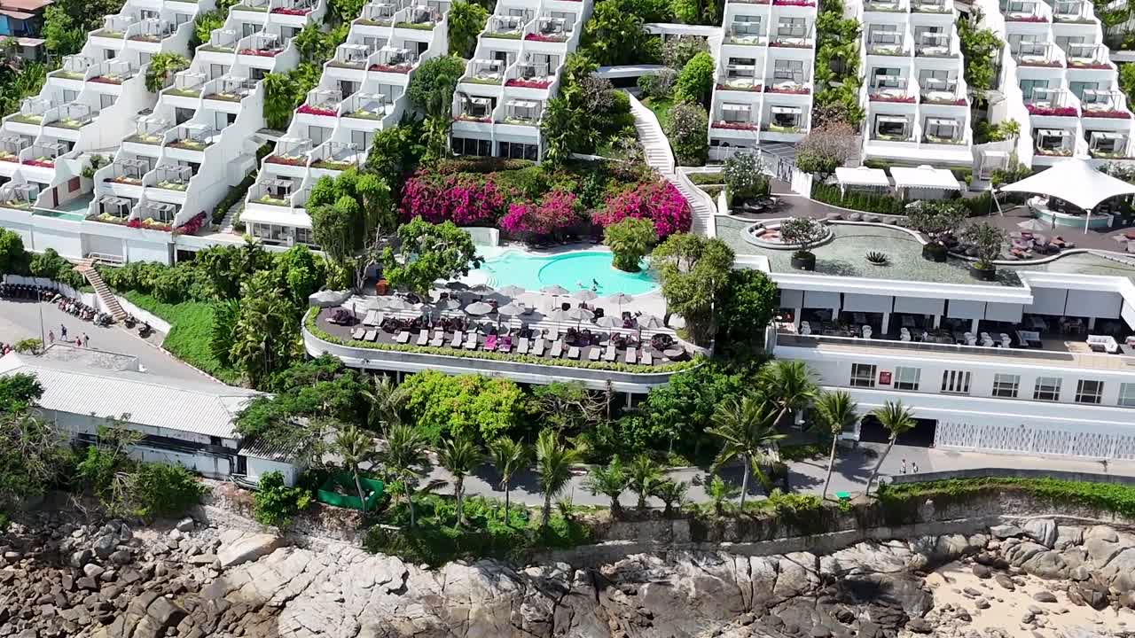 Drone captures a resort with lush gardens and a vibrant beach scene, featuring sunbathers and turquoise waters.