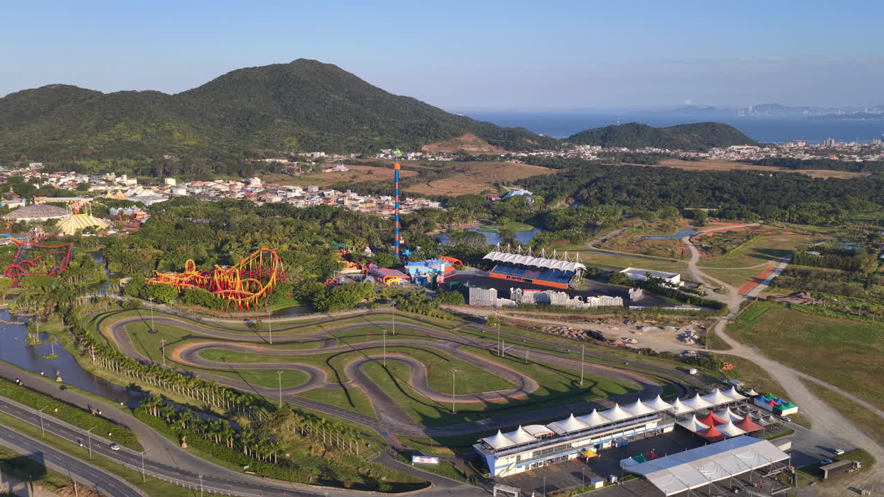 Wide aerial view of Beto Carrero World in Penha, Brazil, showing the winding go-kart racing track, colorful roller coaster, and drop tower framed by lush green hills and coastal surroundings