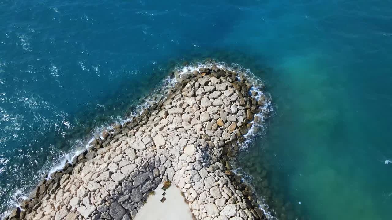 Drone video capturing Durrës breakwater with turquoise waters, showcasing stunning wave patterns. An aerial view of rocky coast under bright lighting and clear skies