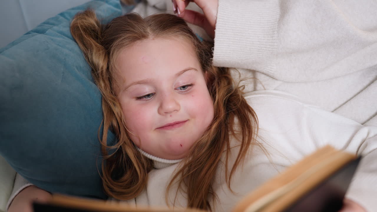 Little girl with long hair lies on bed reading book, face calm and focused, eyes following text, cozy atmosphere with pillow and soft sweater, highlighting childhood curiosity, learning at home