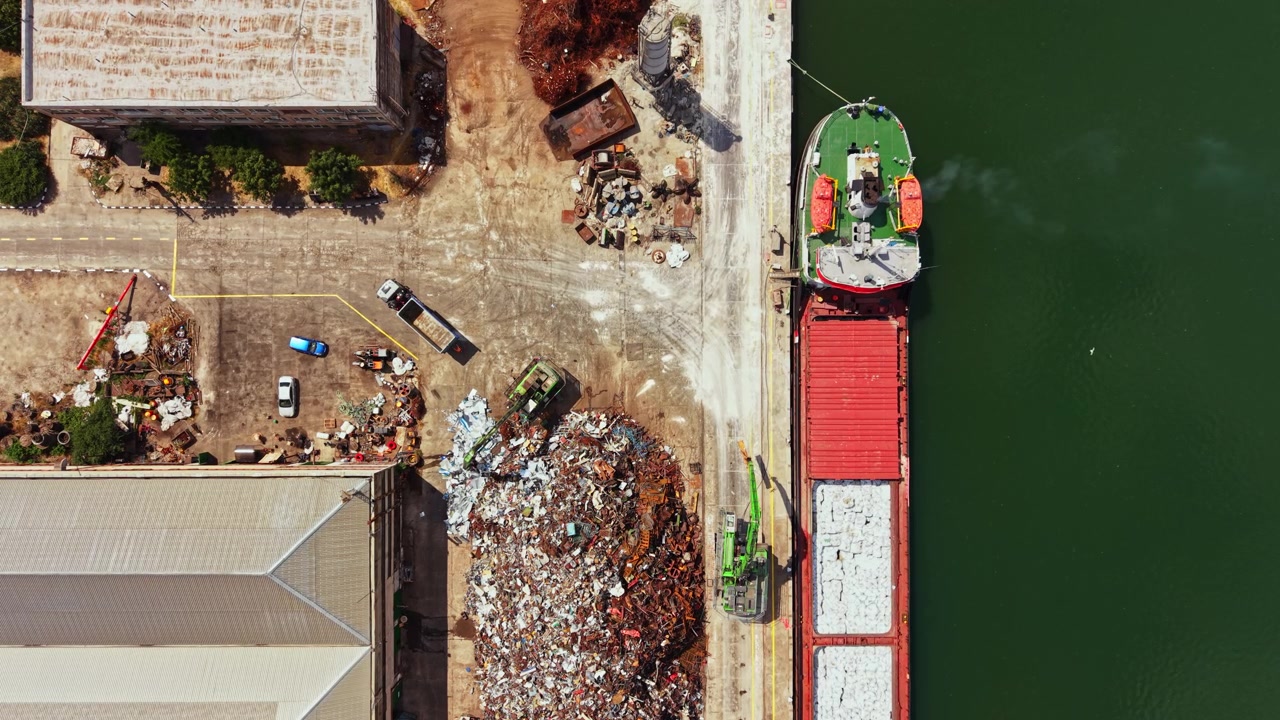 Macchinari pesanti lavorano in un molo dove una grande nave viene caricata con materiali durante il giorno.