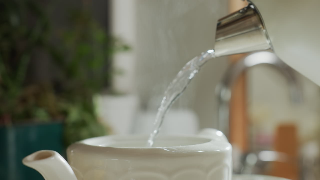 Close up of steam rising from white ceramic mug as hot water is poured into it from kettle with blurred background of warm kitchen setting
