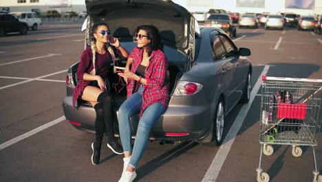 two young attractive women in sunglasses sitting inside of the open car trunk and listening to the music in the smartphone