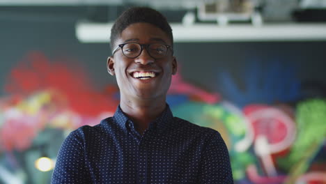 young black male creative smiling to camera in front of mural in casual office, head and shoulders