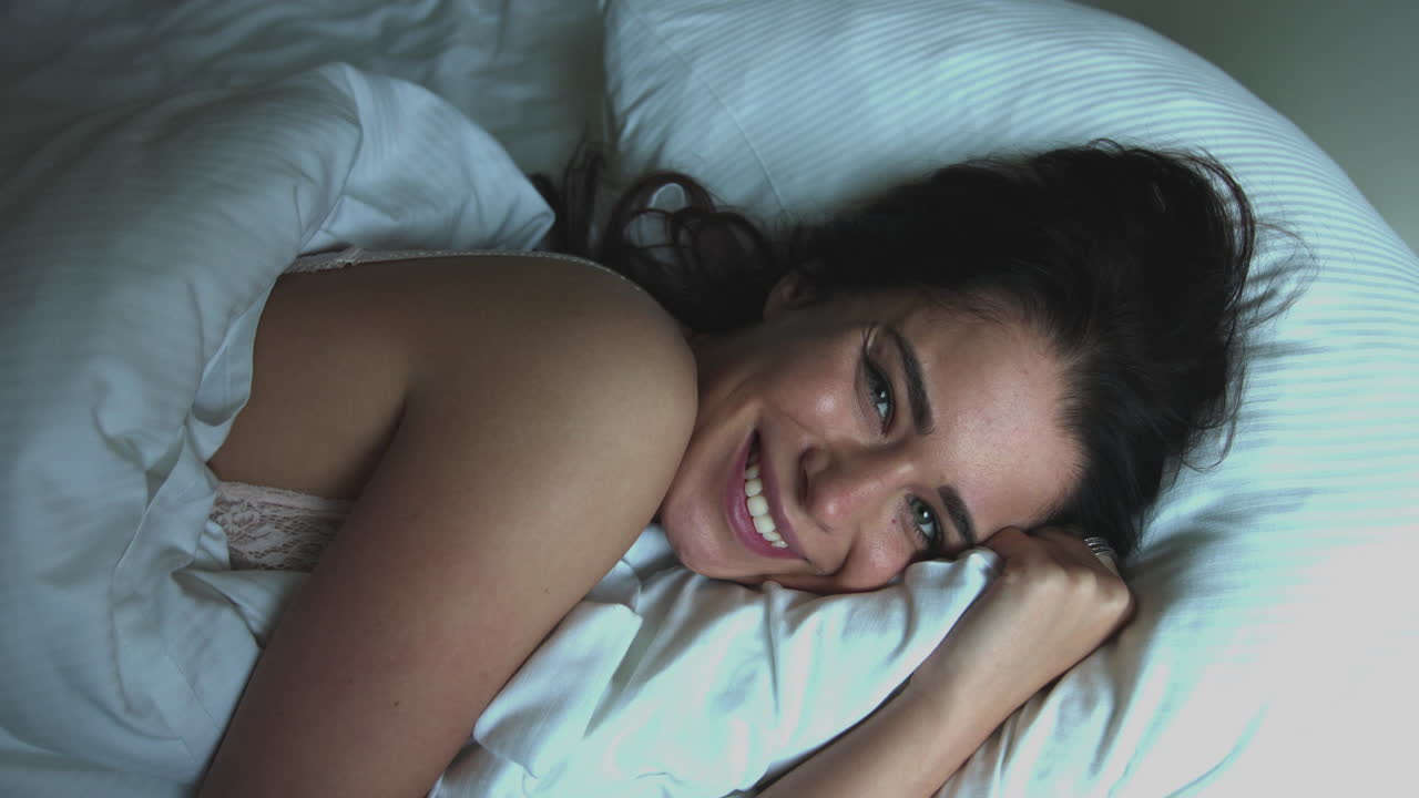 A young woman with a smile on her face lies in bed on a pillow. He is smiling at the camera. A happy morning after a nap. Bed rest