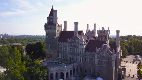 aerial daytime medium shot rising above green trees showing casa loma castle house in summer sunshine with downtown city skyline background in toronto ontario canada