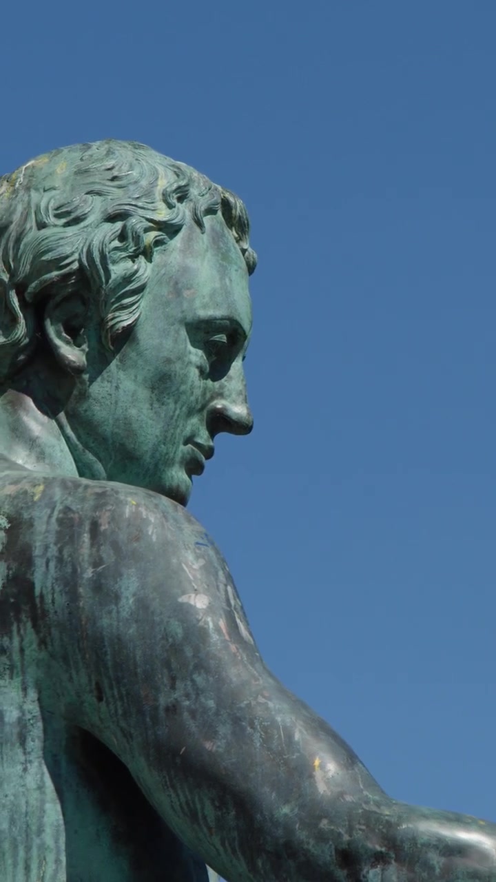 La telecamera esegue una lenta panoramica per rivelare una statua in bronzo di un filosofo contro la guglia di una cattedrale e un cielo azzurro limpido.