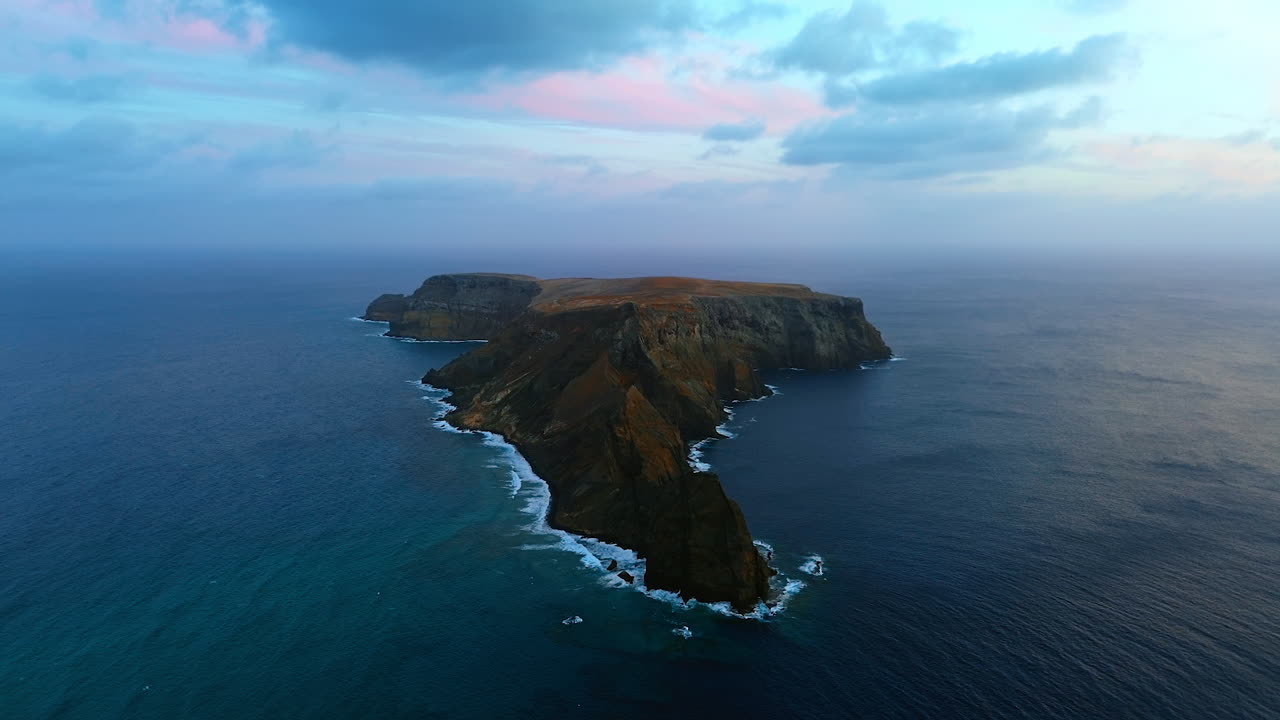 Bare rocky island surrounded by vast waterscape. Grey clouds cover the horizon at backdrop. The Madeira Islands, Portugal. Aerial perspective.