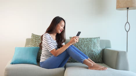 Casual,-attractive-Asian-girl-typing-on-smartphone-while-sitting-on-sofa