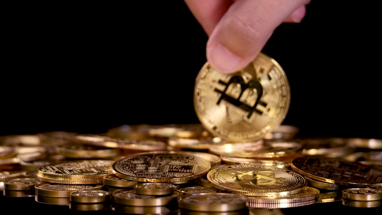 Hand drops golden cryptocurrency coin onto pile of tokens, dramatic lighting, black background, close-up