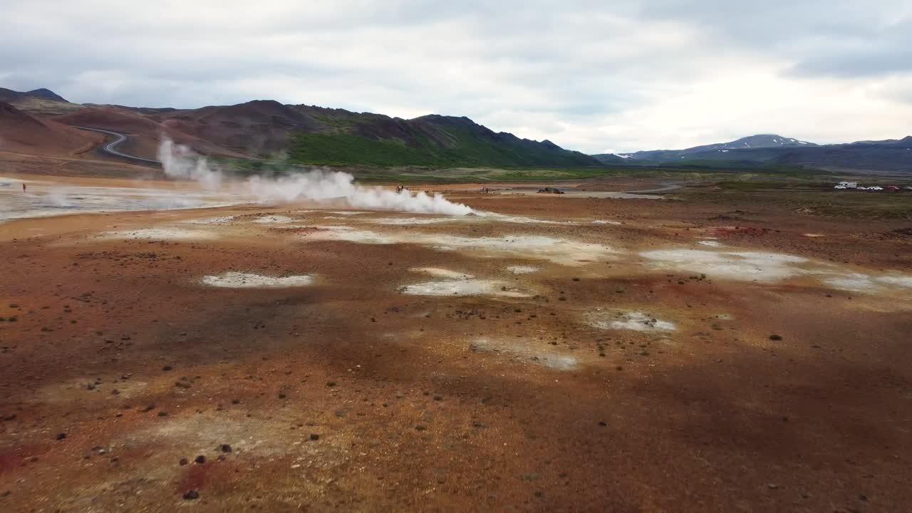 Aerial drone view of steaming geothermal vents in barren volcanic terrain near Hverir, surrounded by colorful mineral deposits, distant hills, and winding road in northern Iceland