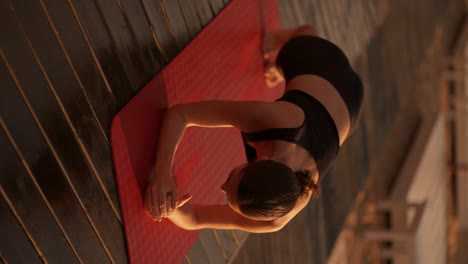 Vertical-video:-a-brunette-girl-in-a-black-tracksuit-does-a-plank-and-periodically-stands-on-her-elbows-on-a-sunny-beach-covered-with-black-boards-in-the-summer