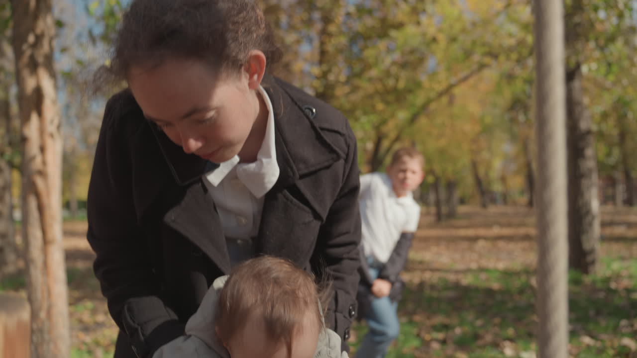 Madre bianca che aiuta il bambino a scendere dall'altalena, primo piano di mani che guidano l'equilibrio, foglie autunnali sotto i piedi, espressione attenta e supporto premuroso, madre come guida paziente che insegna il movimento sicuro.