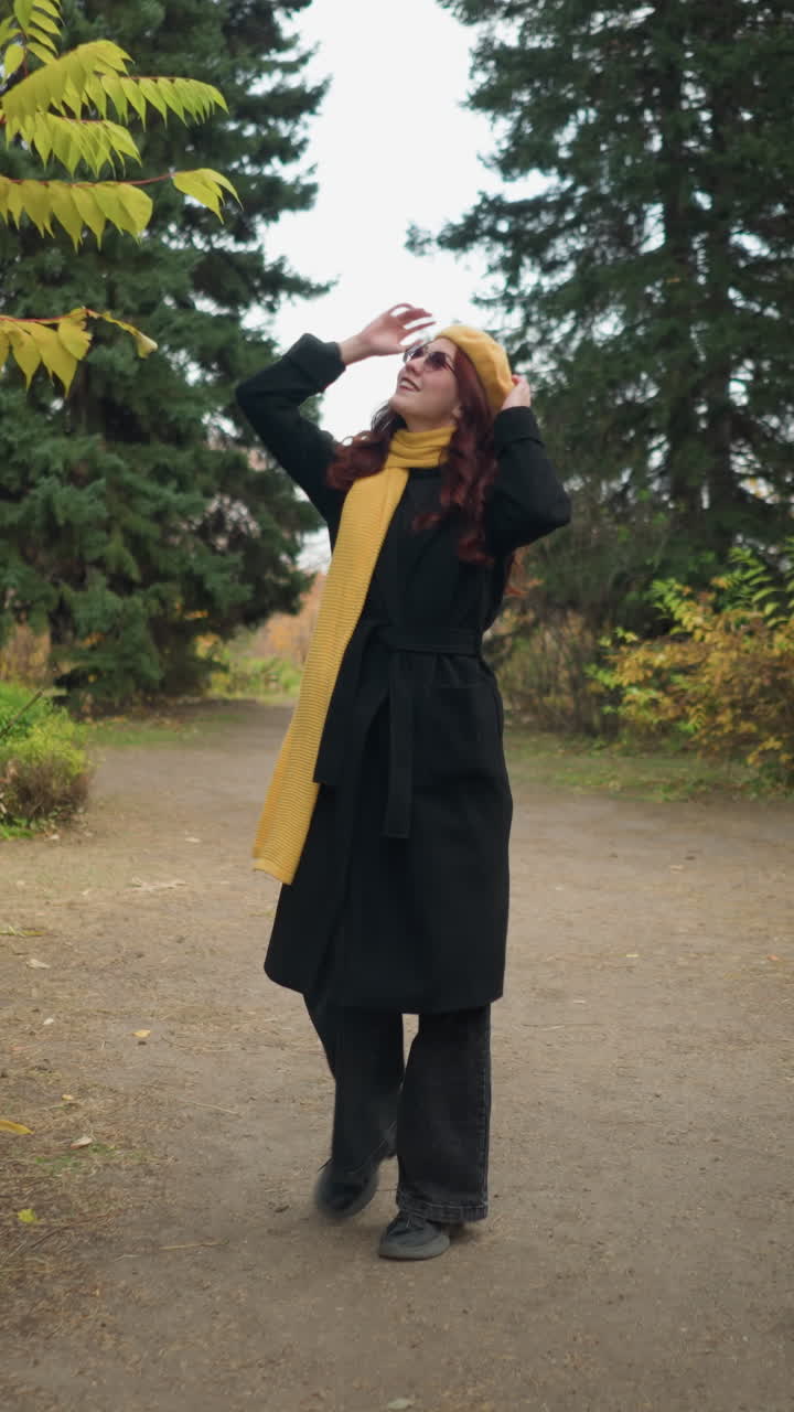 Bella signora con lunghi capelli rossi che indossa un cappotto nero, un basco giallo e una sciarpa, cammina lungo un pittoresco sentiero del giardino circondata da un colorato fogliame autunnale, guardandosi intorno mentre si porta una mano alla testa.