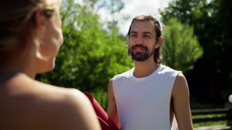 first-person view of a blonde girl in a sports uniform listens to a brunette guy in a white t-shirt who tells her and gesticulates with a yellow bottle in his hands against the backdrop of a green sunny summer park