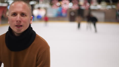 bald man in a brown sweater and black scarf skating at an ice rink, looking at the camera with a subtle smile, with other skaters in the background